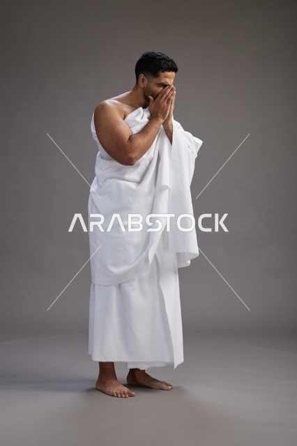 بورتريه لرجل سعودي بملابس الإحرام يعطس ويغطي فمه باحترام, مفهوم الحج والعمره, الجسم بالطول الكامل, خلفيه رماديه