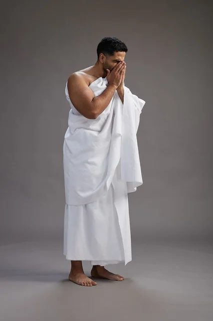 بورتريه لرجل سعودي بملابس الإحرام يعطس ويغطي فمه باحترام, مفهوم الحج والعمره, الجسم بالطول الكامل, خلفيه رماديه