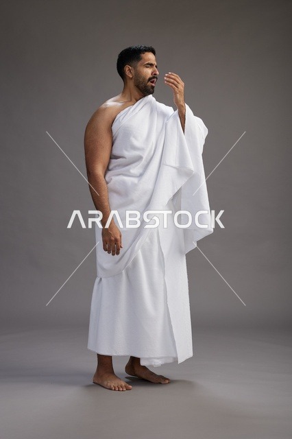 بورتريه لرجل سعودي بملابس الإحرام يعطس ويغطي فمه باحترام, مفهوم الحج والعمره, الجسم بالطول الكامل, خلفيه رماديه