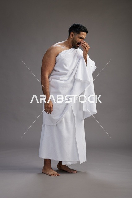 A portrait of a Saudi man in Ihram clothing sneezing and respectfully covering his mouth, the concept of Hajj and Umrah, full-body, gray background.