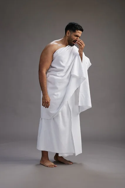 A portrait of a Saudi man in Ihram clothing sneezing and respectfully covering his mouth, the concept of Hajj and Umrah, full-body, gray background. A portrait of a Saudi man in Ihram clothing sneezing and respectfully covering his mouth, the concept of Hajj and Umrah, full-body, gray background.