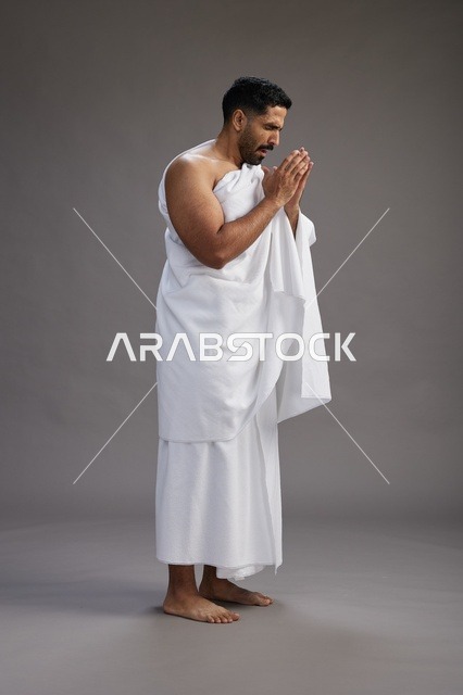 بورتريه لرجل سعودي بملابس الإحرام يعطس ويغطي فمه باحترام, مفهوم الحج والعمره, الجسم بالطول الكامل, خلفيه رماديه