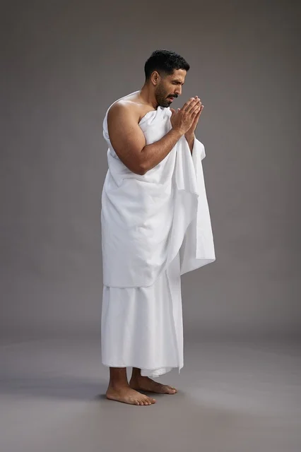 A portrait of a Saudi man in Ihram clothing sneezing and respectfully covering his mouth, the concept of Hajj and Umrah, full-body, gray background.