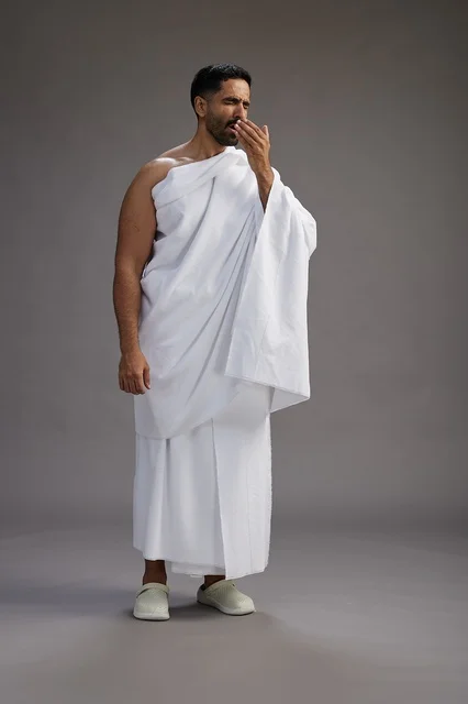 A portrait of a Saudi man in Ihram clothing sneezing and respectfully covering his mouth, the concept of Hajj and Umrah, full-body, gray background.