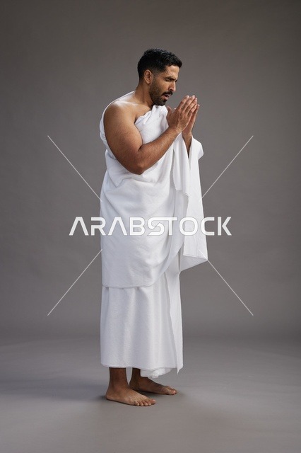 بورتريه لرجل سعودي بملابس الإحرام يعطس ويغطي فمه باحترام, مفهوم الحج والعمره, الجسم بالطول الكامل, خلفيه رماديه