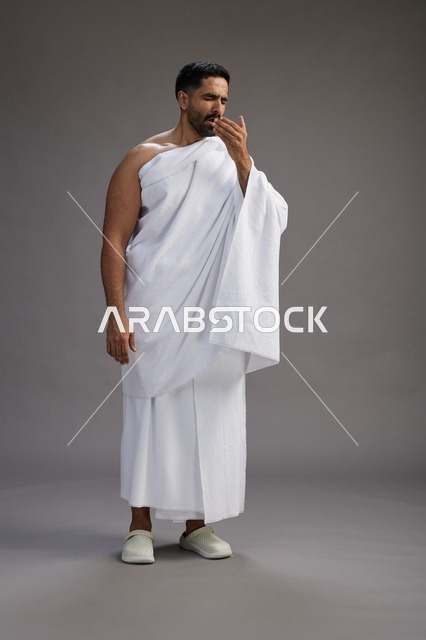 A portrait of a Saudi man in Ihram clothing sneezing and respectfully covering his mouth, the concept of Hajj and Umrah, full-body, gray background.