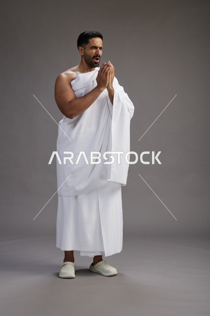 بورتريه لرجل سعودي بملابس الإحرام يعطس ويغطي فمه باحترام, مفهوم الحج والعمره, الجسم بالطول الكامل, خلفيه رماديه