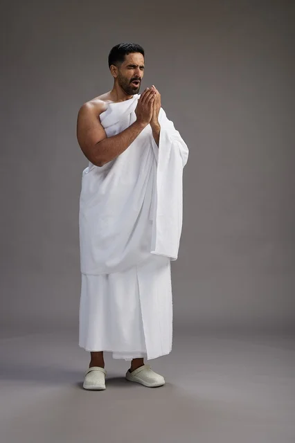 بورتريه لرجل سعودي بملابس الإحرام يعطس ويغطي فمه باحترام, مفهوم الحج والعمره, الجسم بالطول الكامل, خلفيه رماديه