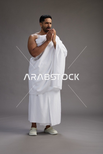 بورتريه لرجل سعودي بملابس الإحرام يعطس ويغطي فمه باحترام, مفهوم الحج والعمره, الجسم بالطول الكامل, خلفيه رماديه