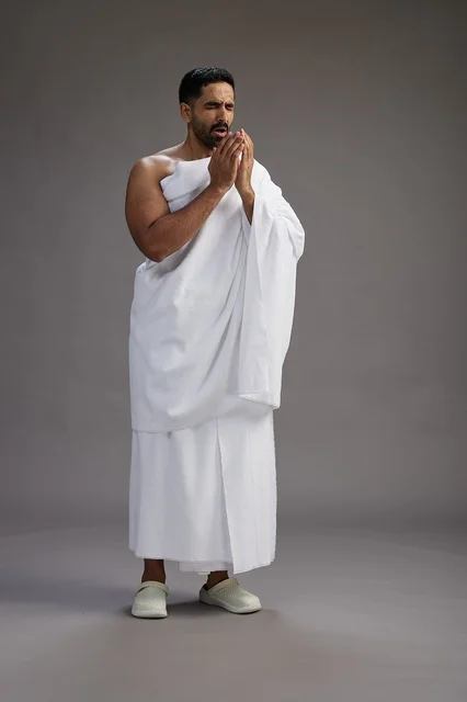 بورتريه لرجل سعودي بملابس الإحرام يعطس ويغطي فمه باحترام, مفهوم الحج والعمره, الجسم بالطول الكامل, خلفيه رماديه