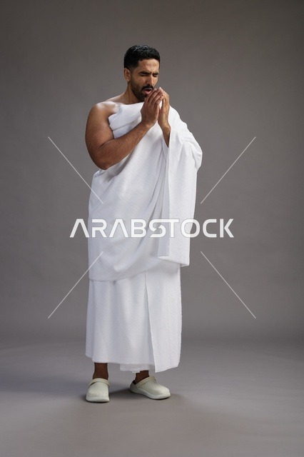 بورتريه لرجل سعودي بملابس الإحرام يعطس ويغطي فمه باحترام, مفهوم الحج والعمره, الجسم بالطول الكامل, خلفيه رماديه
