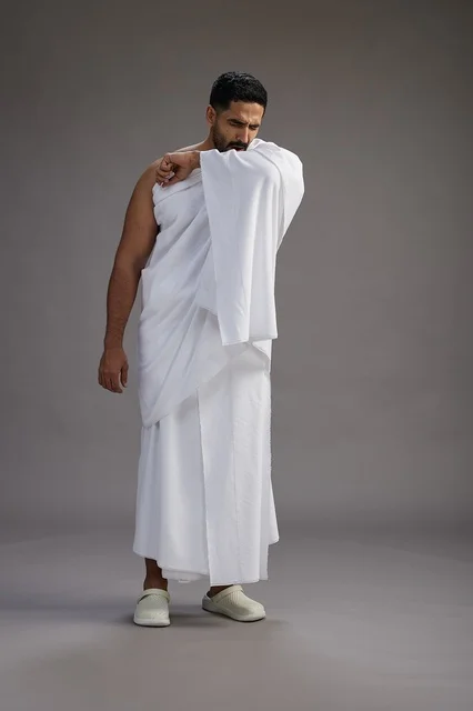 A portrait of a Saudi man in Ihram clothing sneezing and respectfully covering his mouth, the concept of Hajj and Umrah, full-body, gray background.