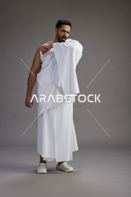 بورتريه لرجل سعودي بملابس الإحرام يعطس ويغطي فمه باحترام, مفهوم الحج والعمره, الجسم بالطول الكامل, خلفيه رماديه