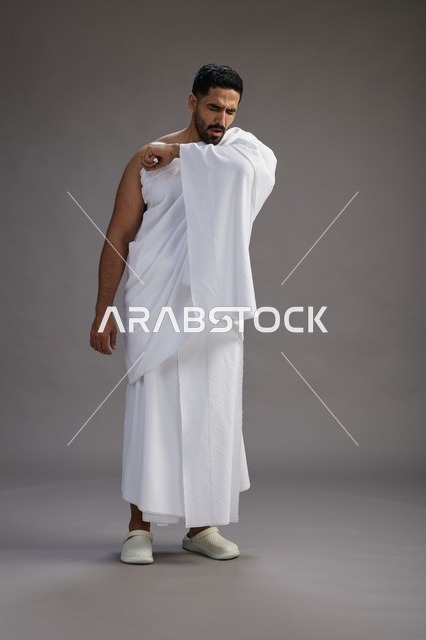 بورتريه لرجل سعودي بملابس الإحرام يعطس ويغطي فمه باحترام, مفهوم الحج والعمره, الجسم بالطول الكامل, خلفيه رماديه