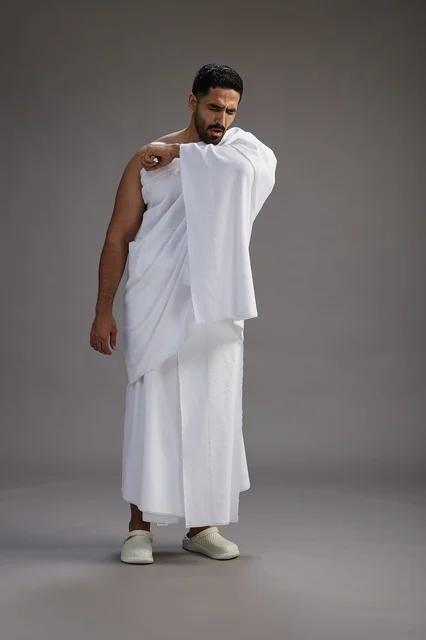 بورتريه لرجل سعودي بملابس الإحرام يعطس ويغطي فمه باحترام, مفهوم الحج والعمره, الجسم بالطول الكامل, خلفيه رماديه