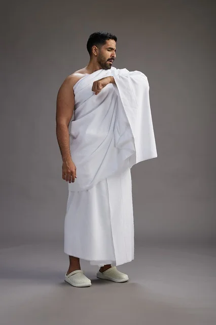 بورتريه لرجل سعودي بملابس الإحرام يعطس ويغطي فمه باحترام, مفهوم الحج والعمره, الجسم بالطول الكامل, خلفيه رماديه