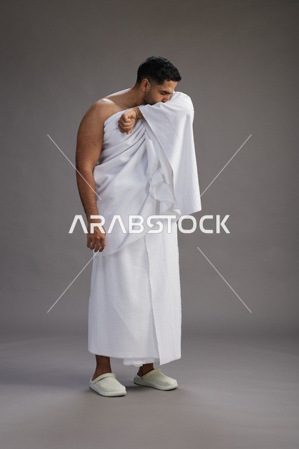 A portrait of a Saudi man in Ihram clothing sneezing and respectfully covering his mouth, the concept of Hajj and Umrah, full-body, gray background.