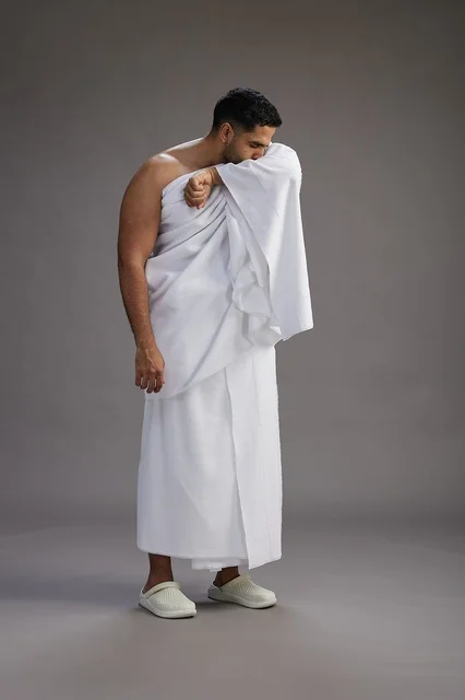 بورتريه لرجل سعودي بملابس الإحرام يعطس ويغطي فمه باحترام, مفهوم الحج والعمره, الجسم بالطول الكامل, خلفيه رماديه