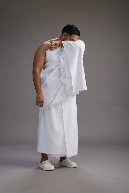 A portrait of a Saudi man in Ihram clothing sneezing and respectfully covering his mouth, the concept of Hajj and Umrah, full-body, gray background. A portrait of a Saudi man in Ihram clothing sneezing and respectfully covering his mouth, the concept of Hajj and Umrah, full-body, gray background.
