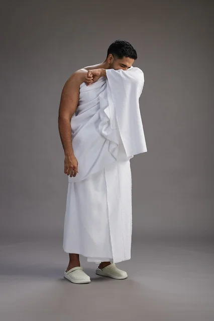 A portrait of a Saudi man in Ihram clothing sneezing and respectfully covering his mouth, the concept of Hajj and Umrah, full-body, gray background. A portrait of a Saudi man in Ihram clothing sneezing and respectfully covering his mouth, the concept of Hajj and Umrah, full-body, gray background.