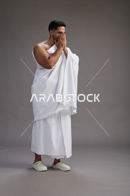 A portrait of a Saudi man in Ihram clothing sneezing and respectfully covering his mouth, the concept of Hajj and Umrah, full-body, gray background.