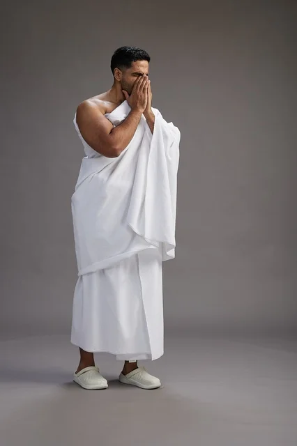 A portrait of a Saudi man in Ihram clothing sneezing and respectfully covering his mouth, the concept of Hajj and Umrah, full-body, gray background.