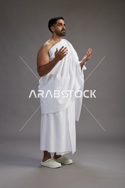 A portrait of a Saudi man in Ihram clothing, praying with his hands raised, supplicating and beseeching God, full body, gray background.