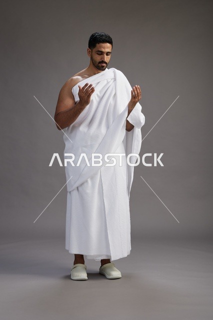 بورتريه لرجل سعودي بملابس الإحرام يدعو رافعاً يديه, الدعاء والتضرع لله , كامل الجسم, خلفيه رماديه