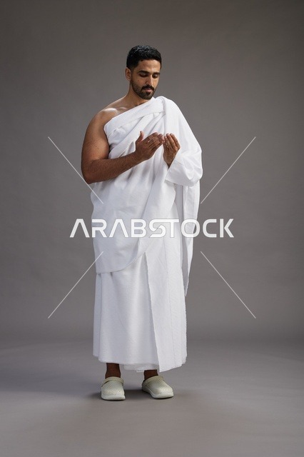 A portrait of a Saudi man in Ihram clothing, praying with his hands raised, supplicating and beseeching God, full body, gray background.