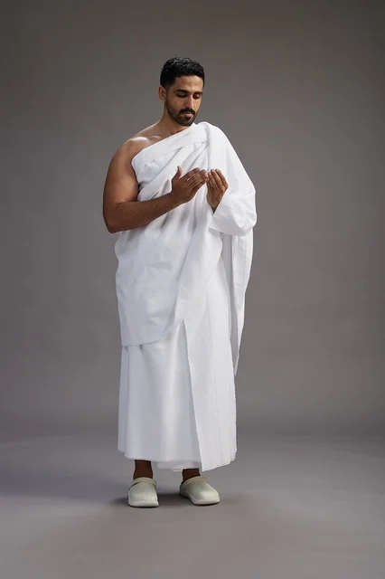 A portrait of a Saudi man in Ihram clothing, praying with his hands raised, supplicating and beseeching God, full body, gray background.