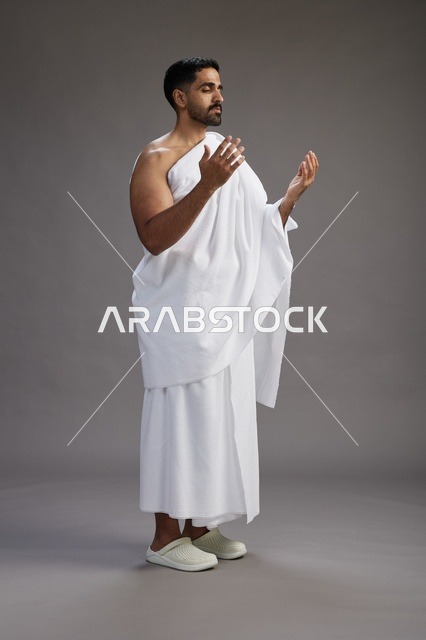 بورتريه لرجل سعودي بملابس الإحرام يدعو رافعاً يديه, الدعاء والتضرع لله , كامل الجسم, خلفيه رماديه