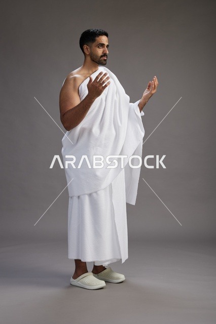 بورتريه لرجل سعودي بملابس الإحرام يدعو رافعاً يديه, الدعاء والتضرع لله , كامل الجسم, خلفيه رماديه