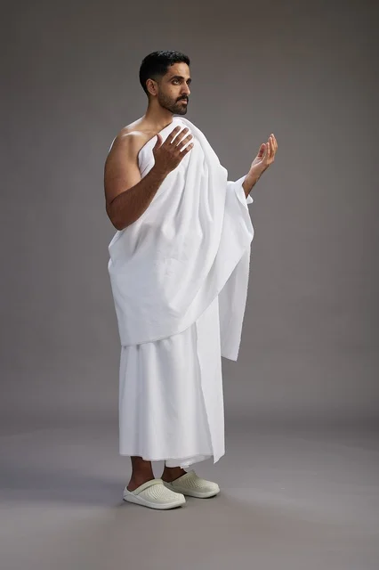 A portrait of a Saudi man in Ihram clothing, praying with his hands raised, supplicating and beseeching God, full body, gray background.