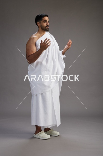 بورتريه لرجل سعودي بملابس الإحرام يدعو رافعاً يديه, الدعاء والتضرع لله , كامل الجسم, خلفيه رماديه