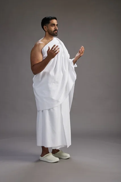 بورتريه لرجل سعودي بملابس الإحرام يدعو رافعاً يديه, الدعاء والتضرع لله , كامل الجسم, خلفيه رماديه