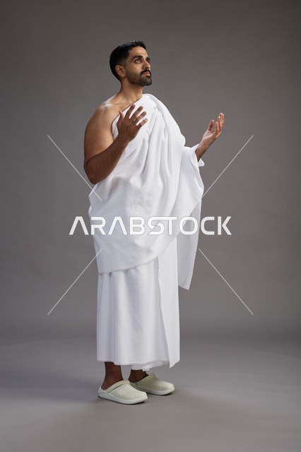 بورتريه لرجل سعودي بملابس الإحرام يدعو رافعاً يديه, الدعاء والتضرع لله , كامل الجسم, خلفيه رماديه