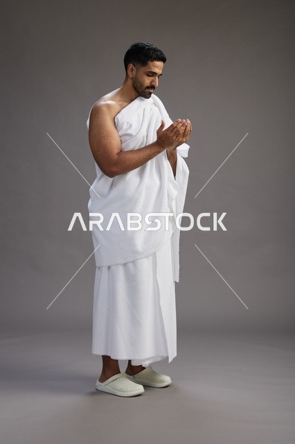 بورتريه لرجل سعودي بملابس الإحرام يدعو رافعاً يديه, الدعاء والتضرع لله , كامل الجسم, خلفيه رماديه