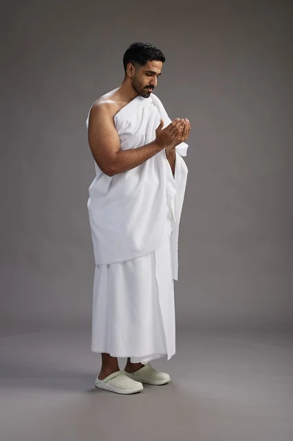 A portrait of a Saudi man in Ihram clothing, praying with his hands raised, supplicating and beseeching God, full body, gray background.