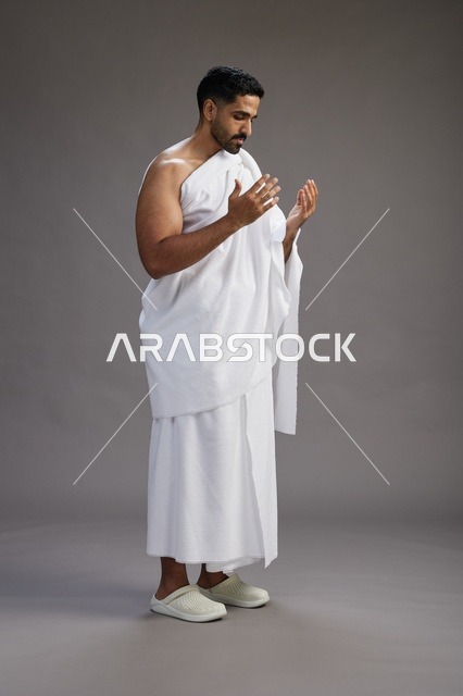 بورتريه لرجل سعودي بملابس الإحرام يدعو رافعاً يديه, الدعاء والتضرع لله , كامل الجسم, خلفيه رماديه