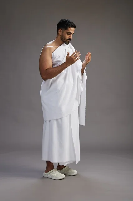 A portrait of a Saudi man in Ihram clothing, praying with his hands raised, supplicating and beseeching God, full body, gray background.