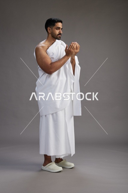 بورتريه لرجل سعودي بملابس الإحرام يدعو رافعاً يديه, الدعاء والتضرع لله , كامل الجسم, خلفيه رماديه
