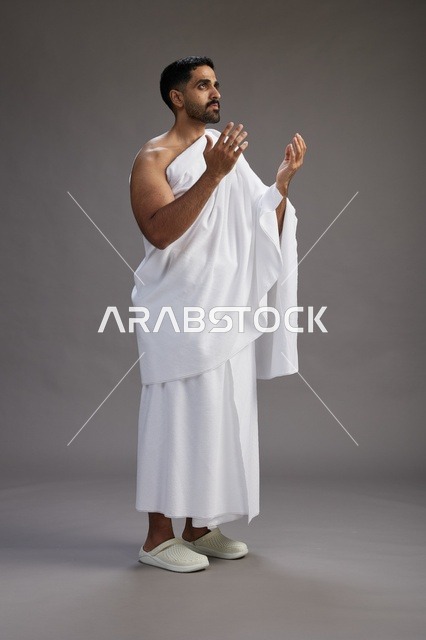 A portrait of a Saudi man in Ihram clothing, praying with his hands raised, supplicating and beseeching God, full body, gray background.