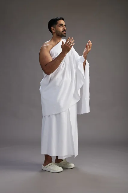 بورتريه لرجل سعودي بملابس الإحرام يدعو رافعاً يديه, الدعاء والتضرع لله , كامل الجسم, خلفيه رماديه