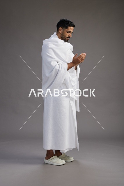 بورتريه لرجل سعودي بملابس الإحرام يدعو رافعاً يديه, الدعاء والتضرع لله , كامل الجسم, خلفيه رماديه