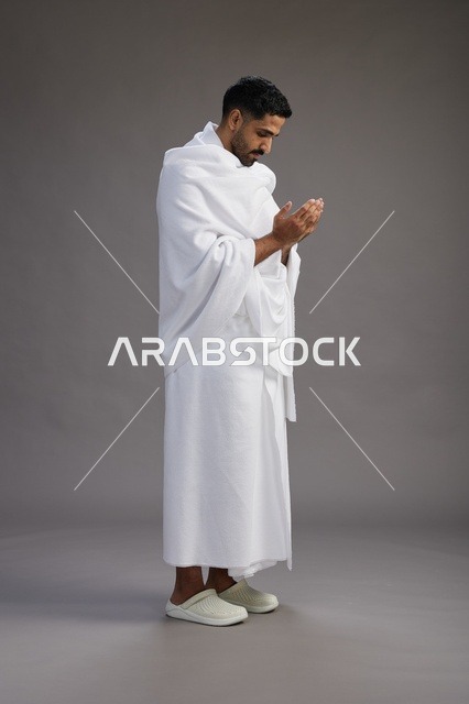 بورتريه لرجل سعودي بملابس الإحرام يدعو رافعاً يديه, الدعاء والتضرع لله , كامل الجسم, خلفيه رماديه