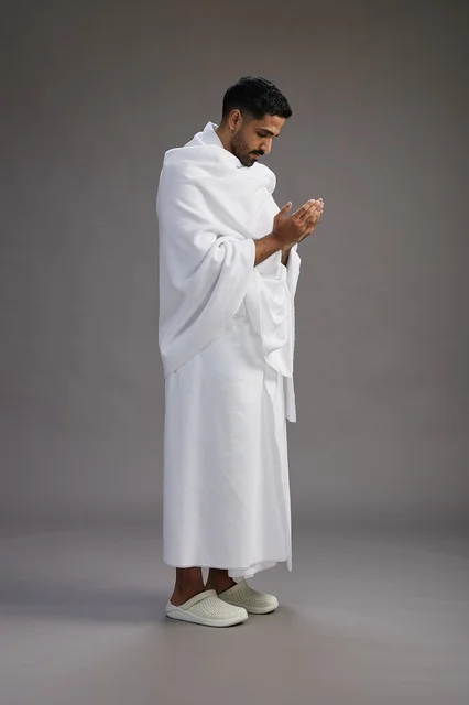 بورتريه لرجل سعودي بملابس الإحرام يدعو رافعاً يديه, الدعاء والتضرع لله , كامل الجسم, خلفيه رماديه