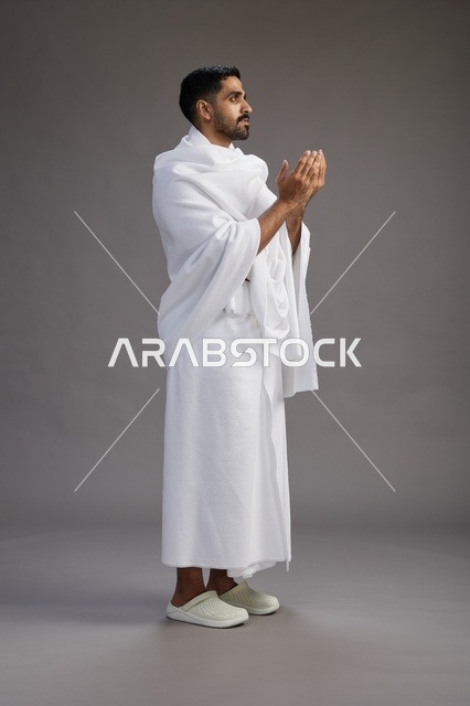 بورتريه لرجل سعودي بملابس الإحرام يدعو رافعاً يديه, الدعاء والتضرع لله , كامل الجسم, خلفيه رماديه