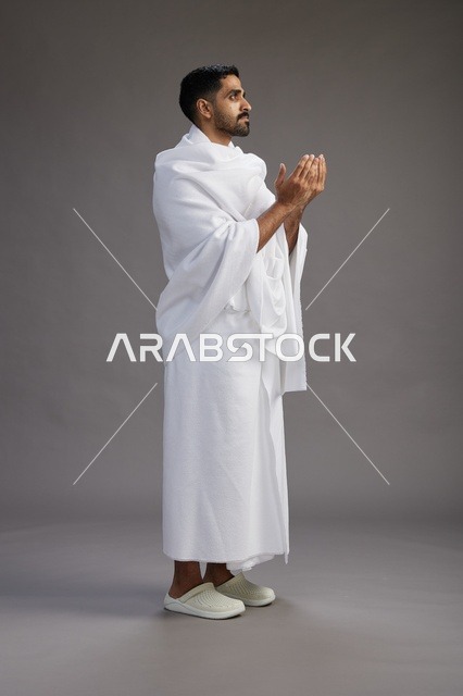 بورتريه لرجل سعودي بملابس الإحرام يدعو رافعاً يديه, الدعاء والتضرع لله , كامل الجسم, خلفيه رماديه