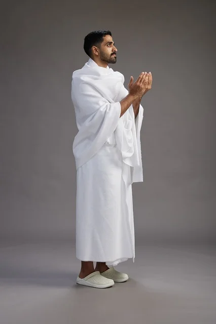 A portrait of a Saudi man in Ihram clothing, praying with his hands raised, supplicating and beseeching God, full body, gray background.