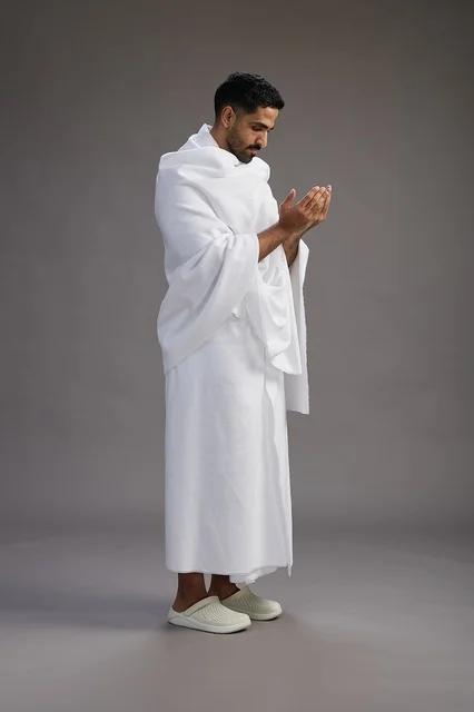 A portrait of a Saudi man in Ihram clothing, praying with his hands raised, supplicating and beseeching God, full body, gray background.